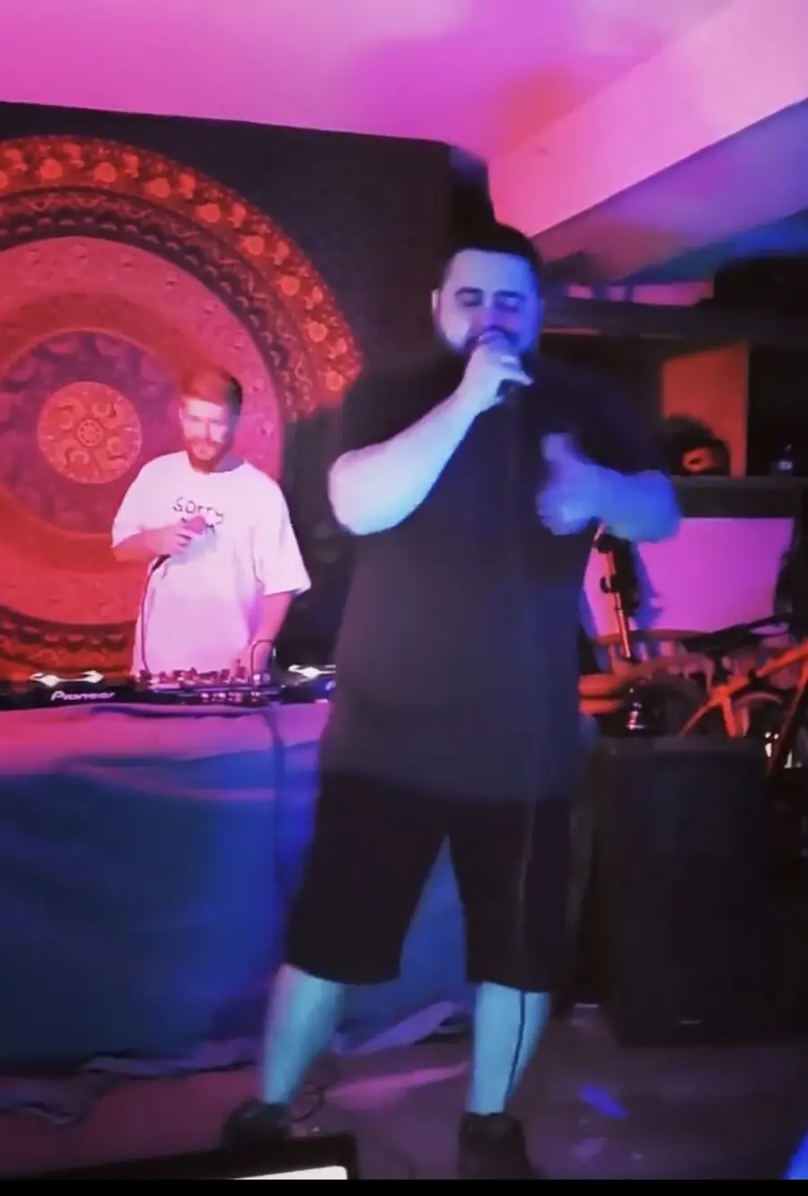 MC Solar Wind rapping into the mic under pink light, Faulkner on the decks behind him